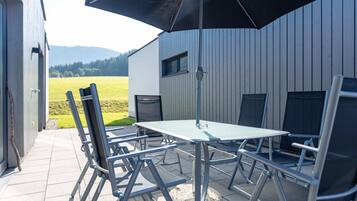 An outdoor patio with a table and chairs, a black umbrella, and a view of a field and mountains in the background.