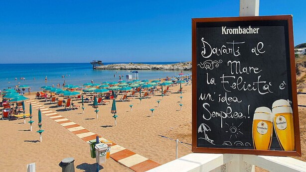 A beach with umbrellas and chairs, a signboard with text, and a view of the sea. - Porticello Sea Village • Three-room apartment (Puglia)