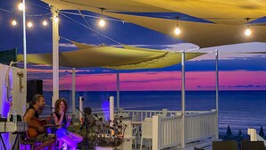 An outdoor dining area with tables and chairs, a stage with a performer, and a view of the ocean. - Porticello Sea Village • Three-room apartment (Puglia)