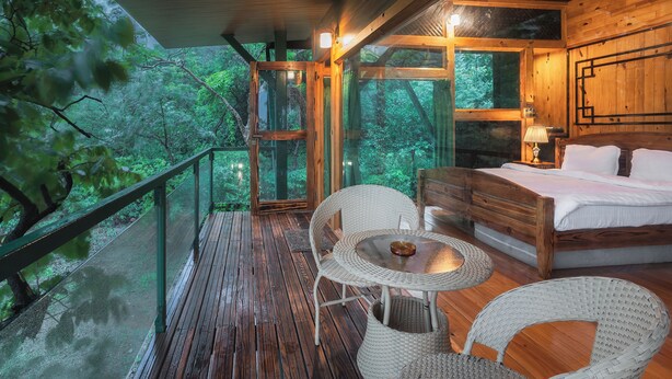 A wooden deck with a glass balustrade overlooking a forest. Inside, there is a bed, a table, and wicker chairs. - The Arboreal Resort (Mawal)