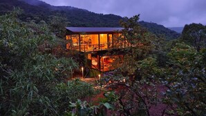 A two-story house with a glass facade, surrounded by dense vegetation and hills in the background. - The Arboreal Resort (Mawal)