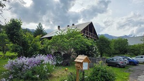 Un edificio rústico con techo inclinado, rodeado de abundante vegetación y plantas en flor.