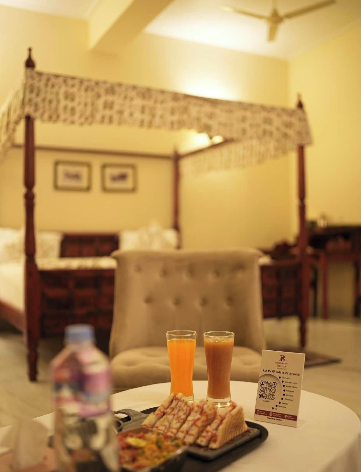 A hotel room with a four-poster bed, a dining table with a tray of food and two glasses of orange juice, and a QR code menu sign.