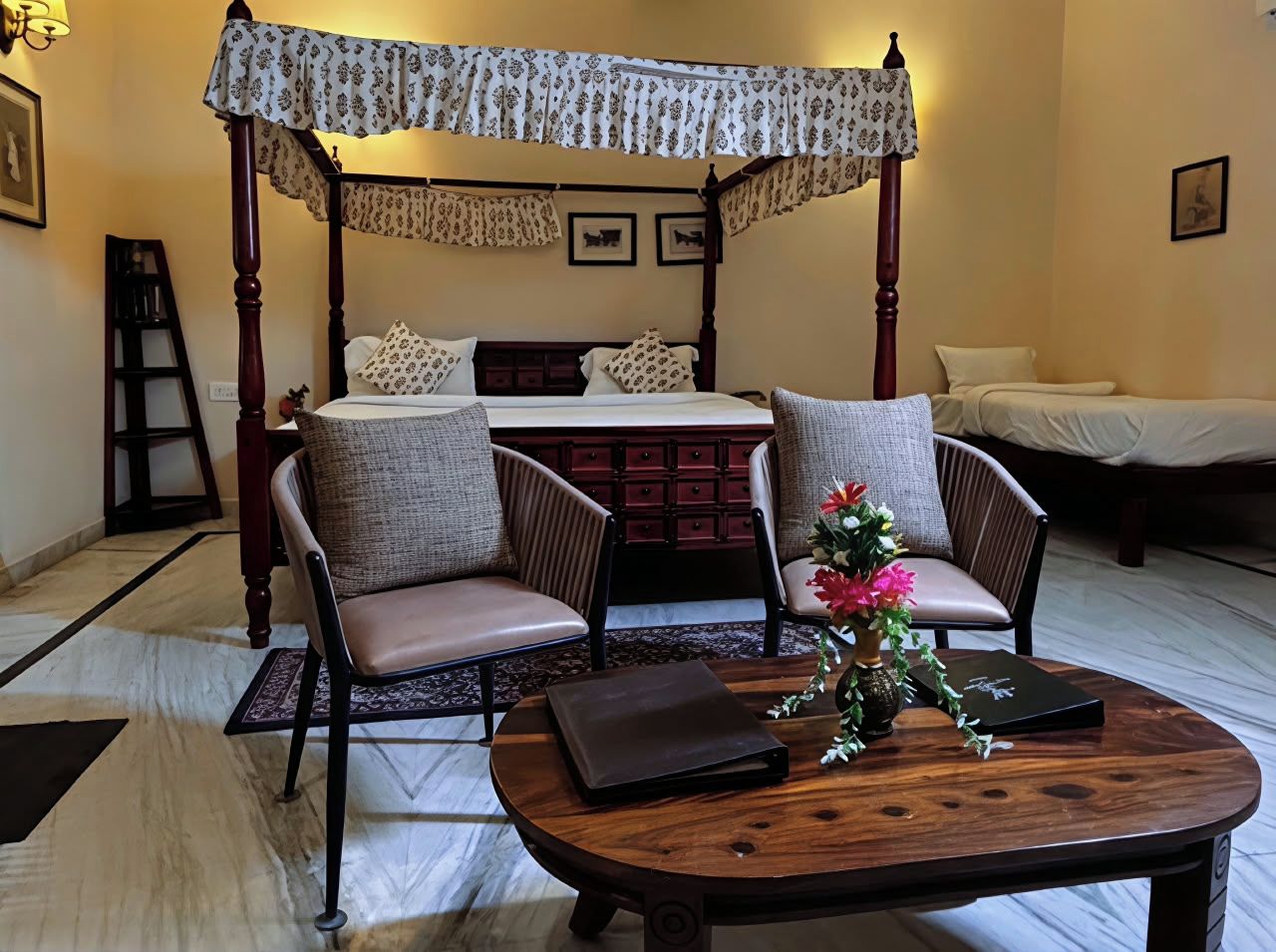 A four-poster bed with a canopy, two upholstered chairs, a small table with a vase, and a nightstand.