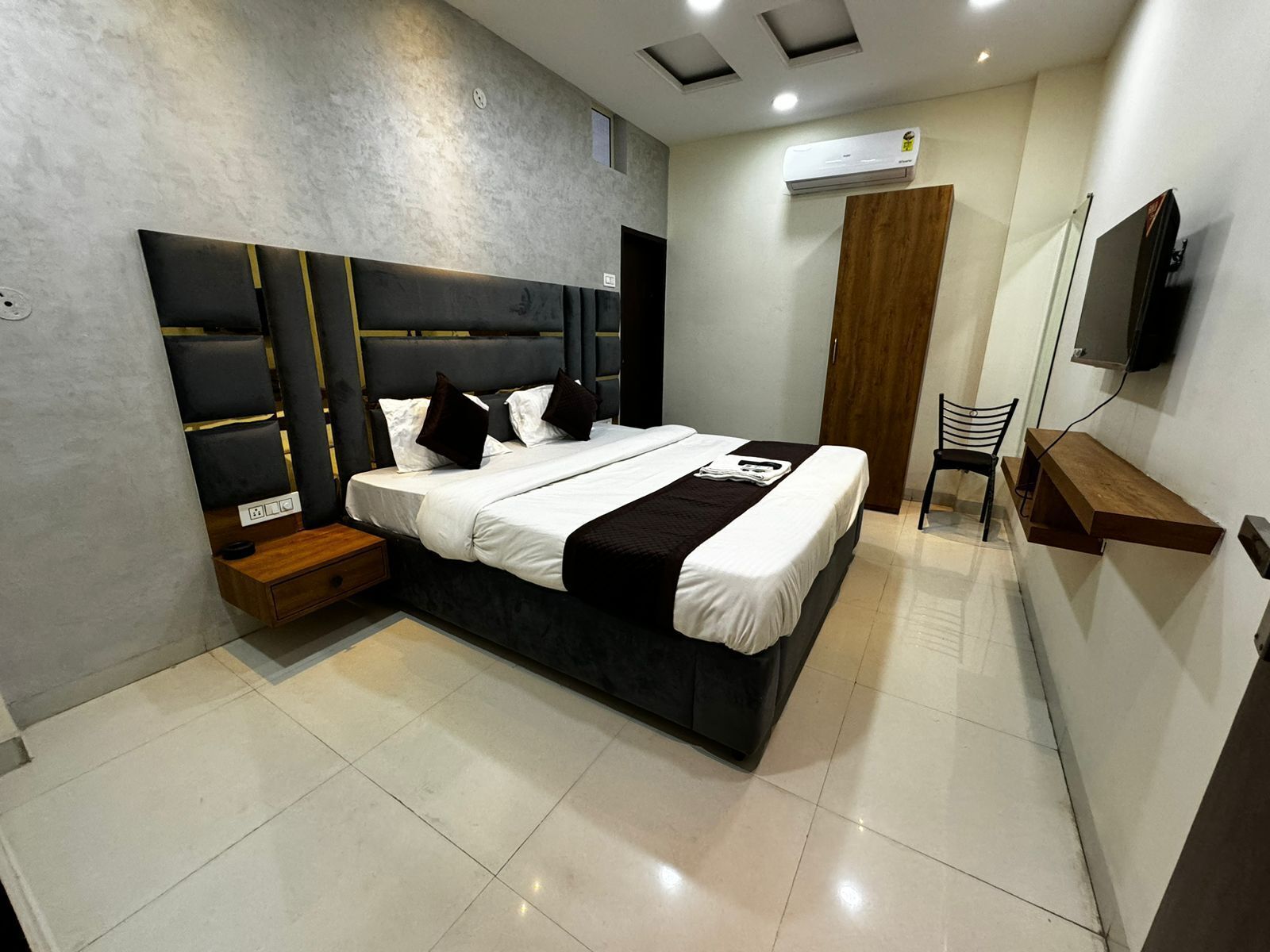 A modern bedroom with a large bed, bedside tables, a wooden chair, a TV, and an air conditioning unit.
