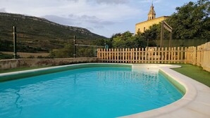 Une piscine entourée d’une clôture en bois, avec vue sur une colline et un bâtiment en arrière-plan.