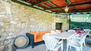 Une terrasse extérieure avec des murs en pierre, un plafond en bois, une table avec des chaises et un coin salon avec des coussins.