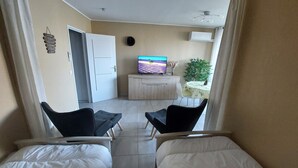A hotel room with two beds, a TV on a wooden cabinet, a dining table with chairs, and a plant.