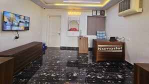A reception area with a reception desk labeled "Namaste!", a ceiling fan, a wall-mounted TV displaying security footage, and a decorative wall piece. - HOTEL AVAAS TripandTrip (Puri)