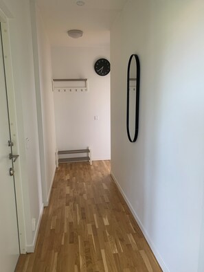 A hallway with a wooden floor, a white door, a black clock, and a wall-mounted mirror. - Villa Nova (Lund)