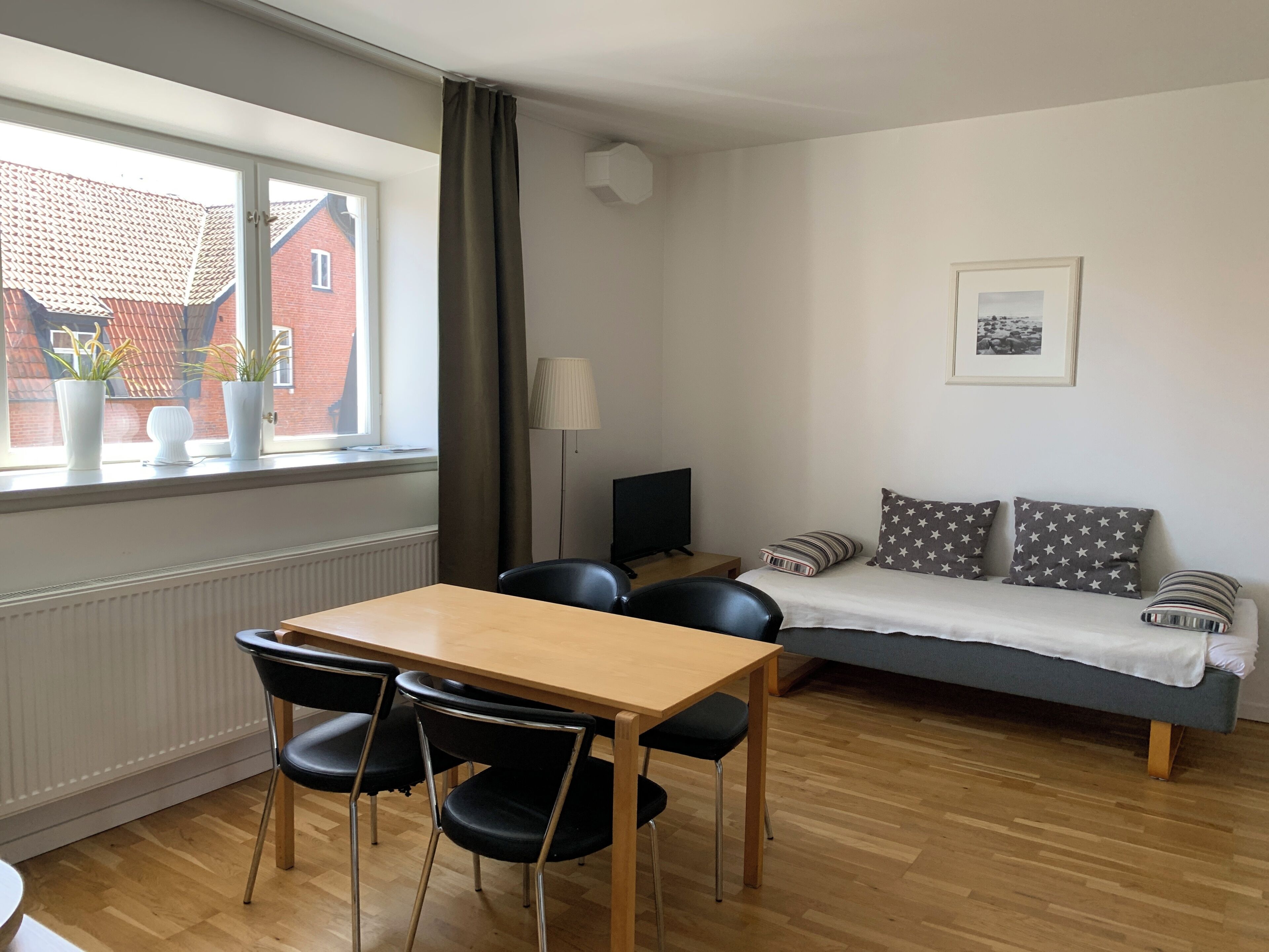 Ein Zimmer mit Holztisch und -stühlen, einem Sofa mit gemusterten Kissen, einem Fenster mit Vorhängen und einer Heizung.