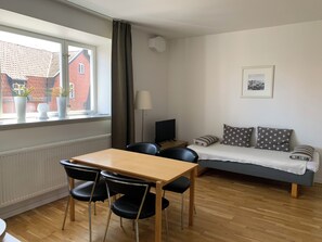A room with a wooden table and chairs, a sofa with patterned cushions, a window with curtains, and a radiator. - Villa Nova (Lund)