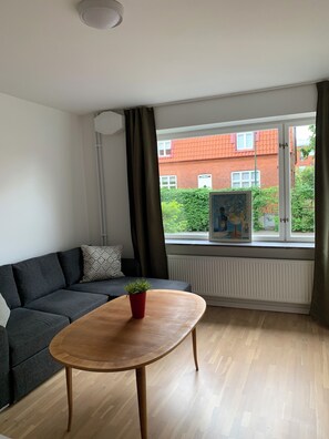A modern living room with a grey sofa, a wooden coffee table with a red pot, and a view of a red brick building through a window with a white radiator below. - Villa Nova (Lund)