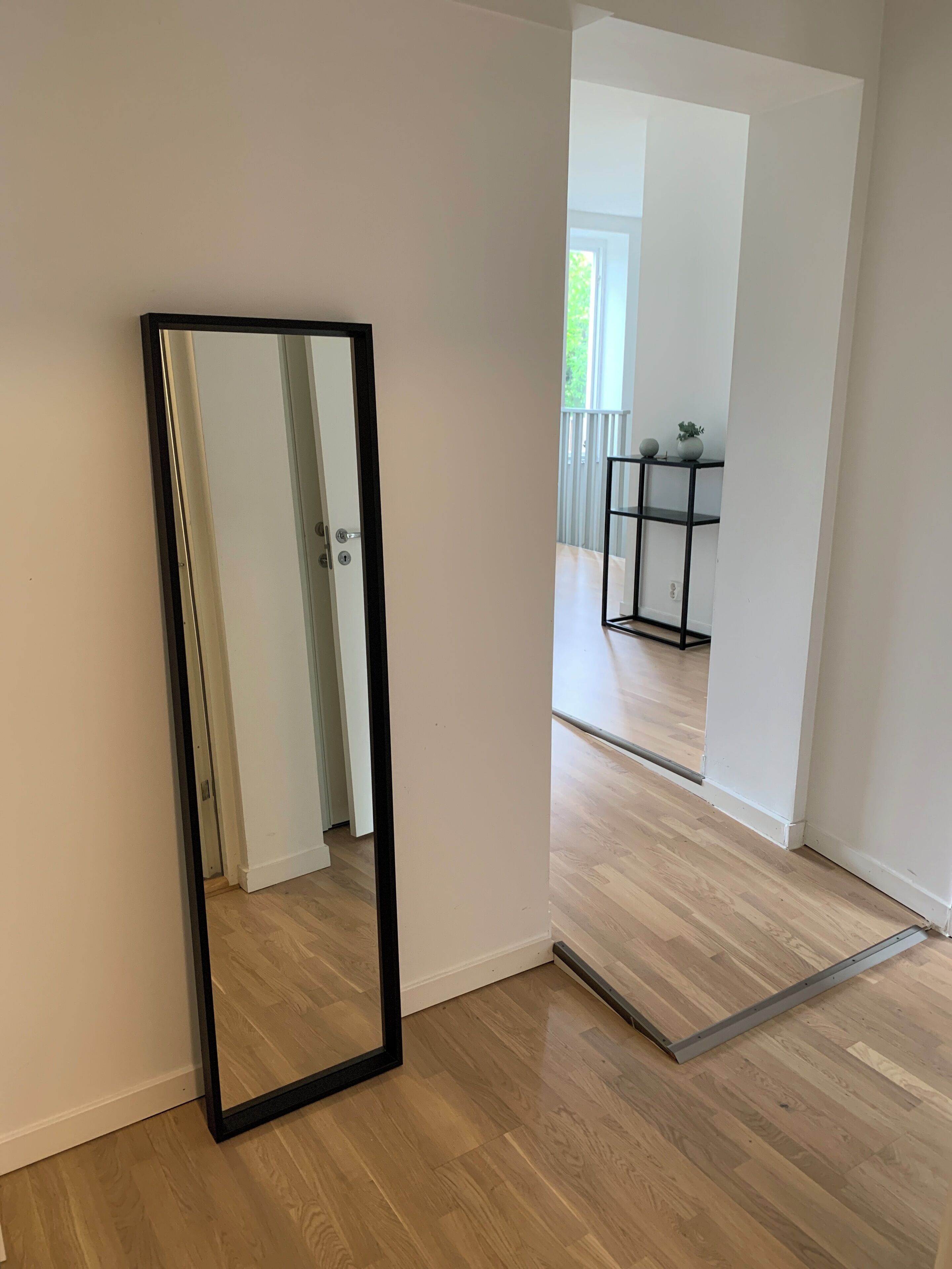 A room with a wooden floor, a black-framed mirror, and a door leading to another room.