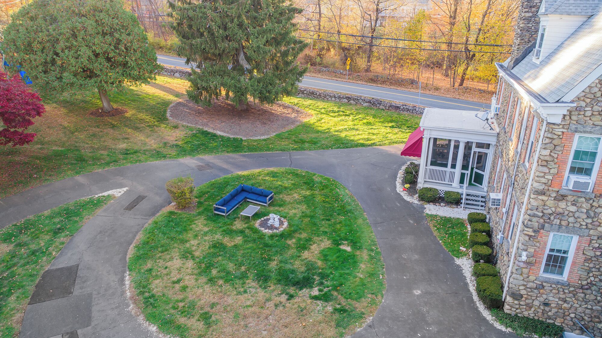A stone house with a porch, a lawn with a blue bench, and a driveway.