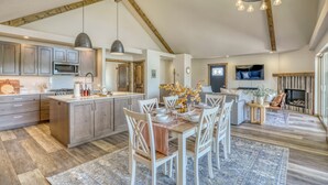 Une cuisine spacieuse avec des poutres en bois, un coin repas avec des chaises blanches et une cheminée dans le salon.