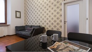A small, modern room with a patterned wallpaper, a grey sofa, a glass table with two mugs, and a door. - Italianway Easy - Plinio 37 (Milan)