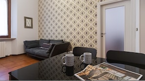A small, modern room with a patterned wallpaper, a grey sofa, a glass table with two mugs, and a door.