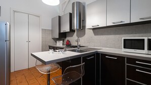 A modern kitchen with a stainless steel refrigerator, a microwave, a sink, and a stove. There is a chair and a table with glasses and a menu. - Italianway Easy - Plinio 37 (Milan)