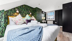 Una habitación de hotel moderna con una cama grande, almohadones estampados con motivos florales, una colcha azul con diseños y una pared con patrón de hojas verdes.