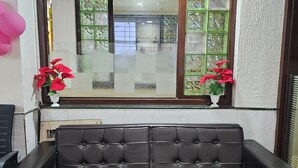 A black leather sofa in a room with a window, two potted red plants, and a wall with a textured pattern. - Hotel Saikrupa (Rahata)