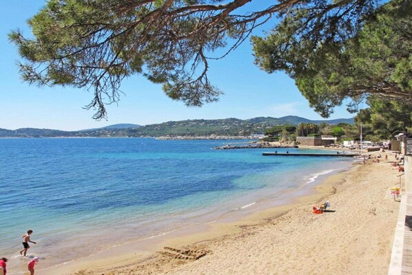 A beach with clear blue water, a sandy shore, and a few people. There are trees and hills in the background.