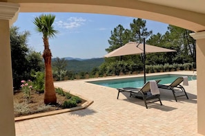A pool area with lounge chairs and a palm tree, surrounded by a paved patio and a view of mountains.