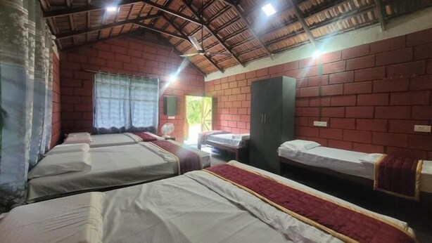 A dormitory room with four single beds, a brick wall, a wooden ceiling, and a window with curtains. - Tusker Valley Resort (Sakleshpur)