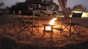 A campsite with a fire pit surrounded by metal chairs, a vehicle in the background, and a tree providing shade. - Moremi Enclave Resort  (Maun)