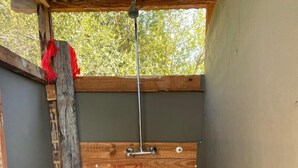 A small, enclosed shower area with a wooden bench, a metal showerhead, and a view of greenery outside. - Moremi Enclave Resort  (Maun)