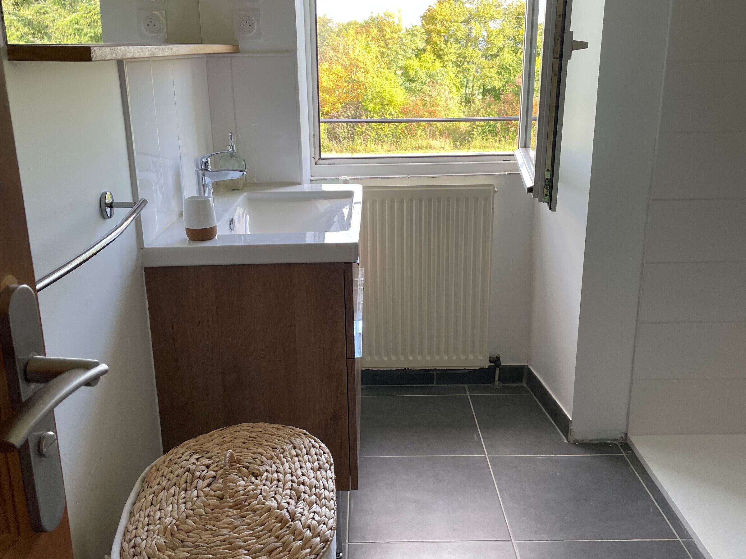 A small bathroom with a window, a wooden cabinet, a sink, and a woven stool.