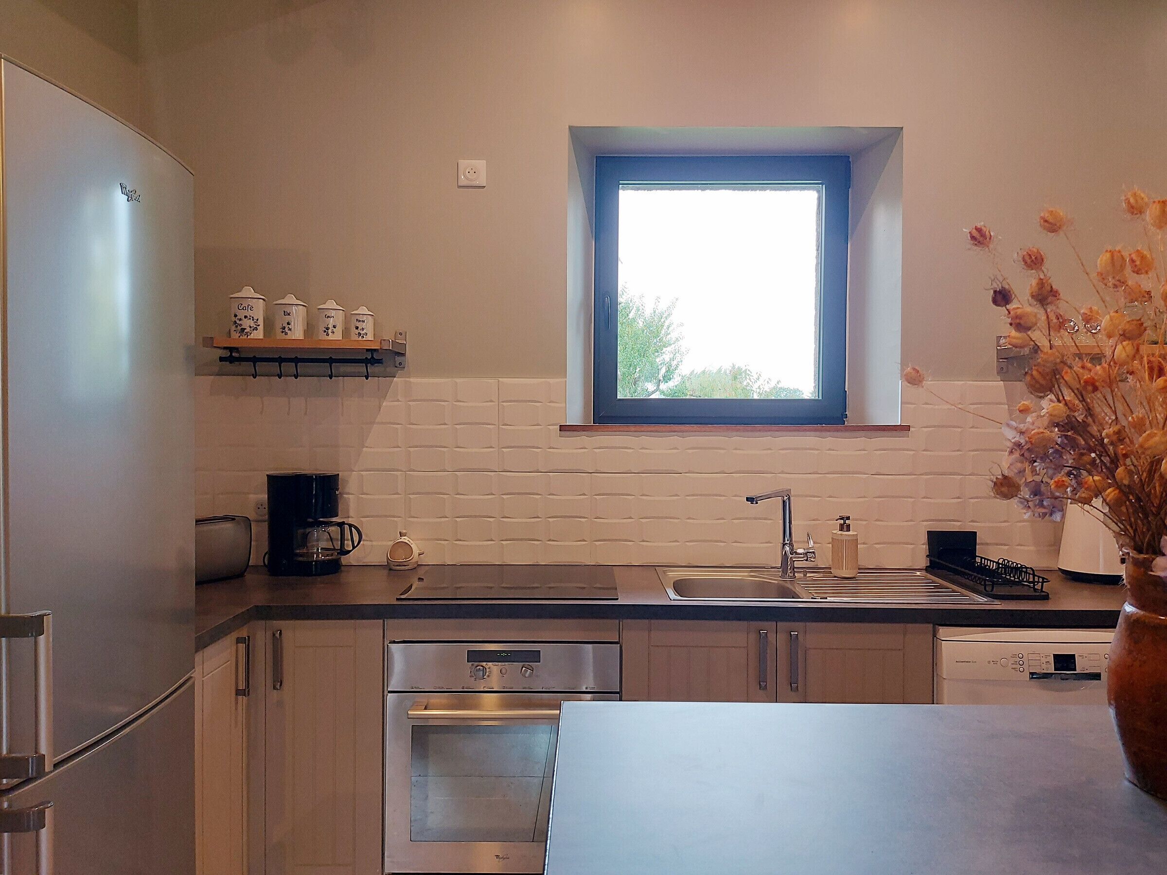 Una cocina moderna con electrodomésticos de acero inoxidable, una ventana sobre el fregadero y un jarrón con flores secas en la encimera.