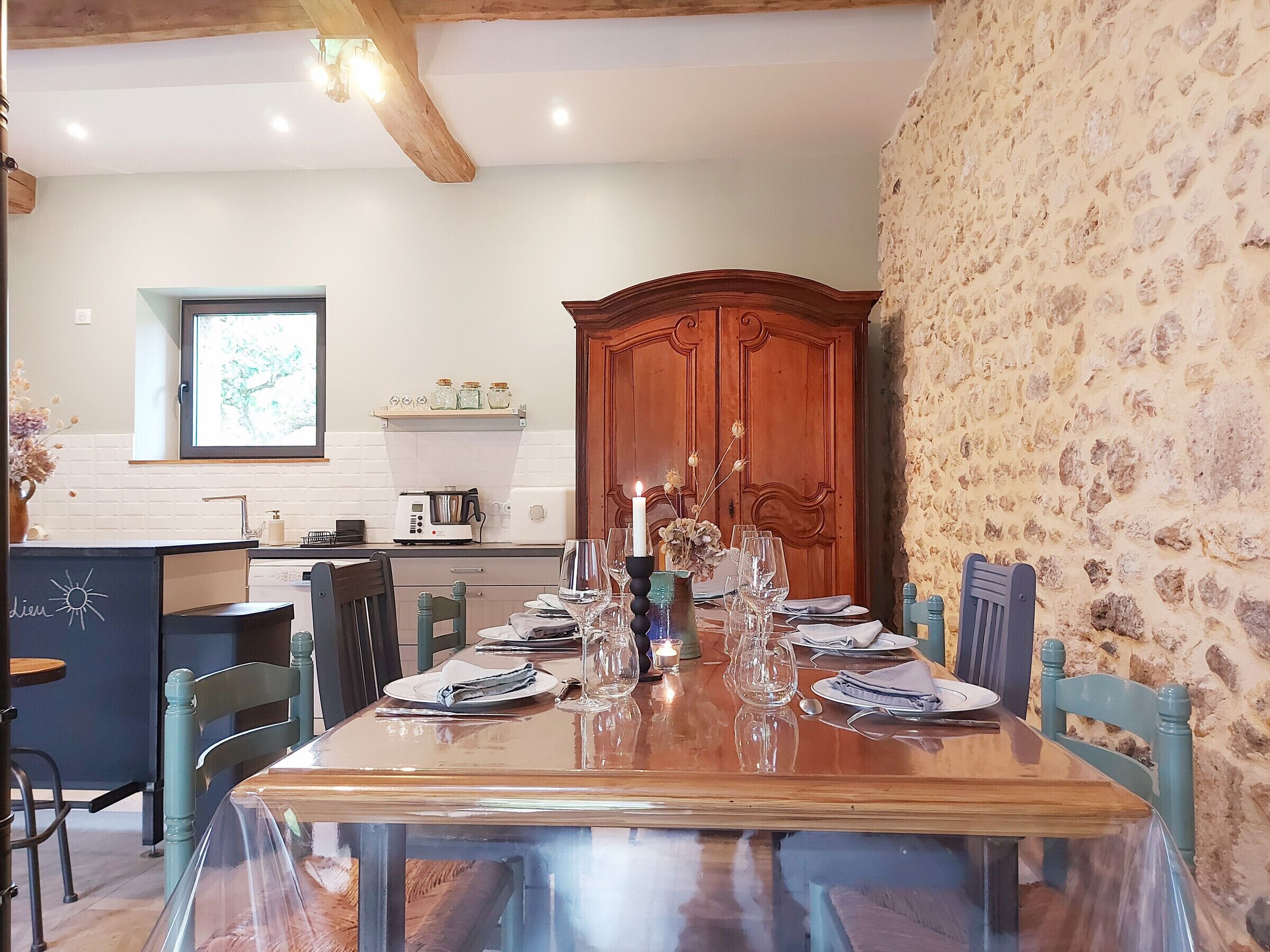 A dining area with a wooden table set for four, a kitchen in the background, and a stone wall.