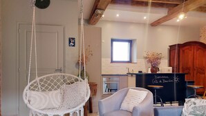 Une pièce avec un fauteuil suspendu en macramé, un fauteuil gris et un canapé bleu. On y trouve également un coin cuisine avec un comptoir noir et une fenêtre au-dessus.