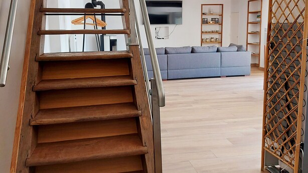 Una scala in legno conduce a un soggiorno con un divano grigio, una TV e scaffali con oggetti.