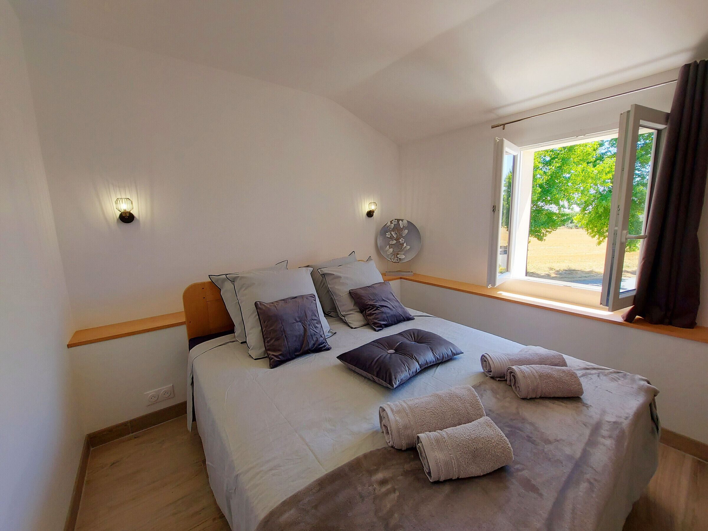 A bedroom with a bed, pillows, a bedside table, a wall clock, and a window with curtains.