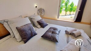 Una camera da letto con un letto, cuscini, un comodino, una lampada a parete e una finestra vista alberi.