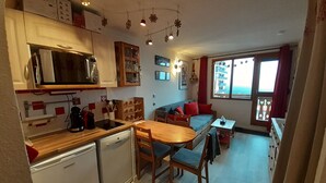 A compact kitchen with a microwave, stove, and small refrigerator. There is a dining area with a table and chairs, and a sofa with red cushions. The room has a large window with curtains and a view of buildings.