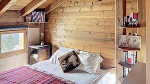 Tunnelmallinen makuuhuone, jossa on puinen, kalteva kattopinta, valkoisilla lakanoiden peitetty sänky ja punavalkoraidallinen peitto, yöpöytä lampun kanssa sekä kirjoja täynnä oleva kirjahylly ja pieni koriste-esine.