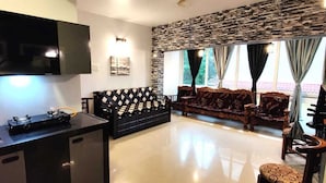 A modern kitchen with a flat-screen TV, a gas stove, and a built-in oven. There is a black sofa with a diamond-patterned cushion, a wooden coffee table, and a stone-textured wall. Large windows with curtains are visible.