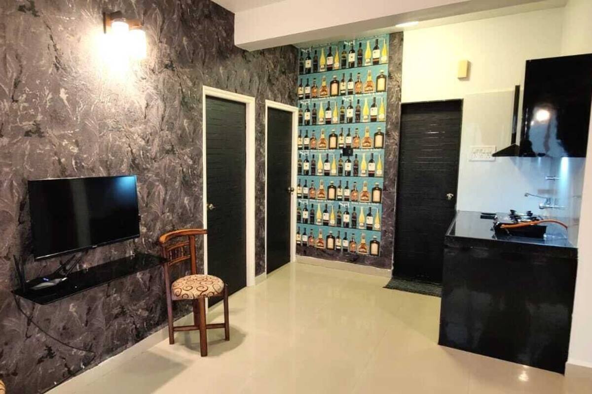 A modern living room with a flat-screen TV, a wooden chair, a wall-mounted shelf with bottles, and a kitchen area with a black countertop and sink.
