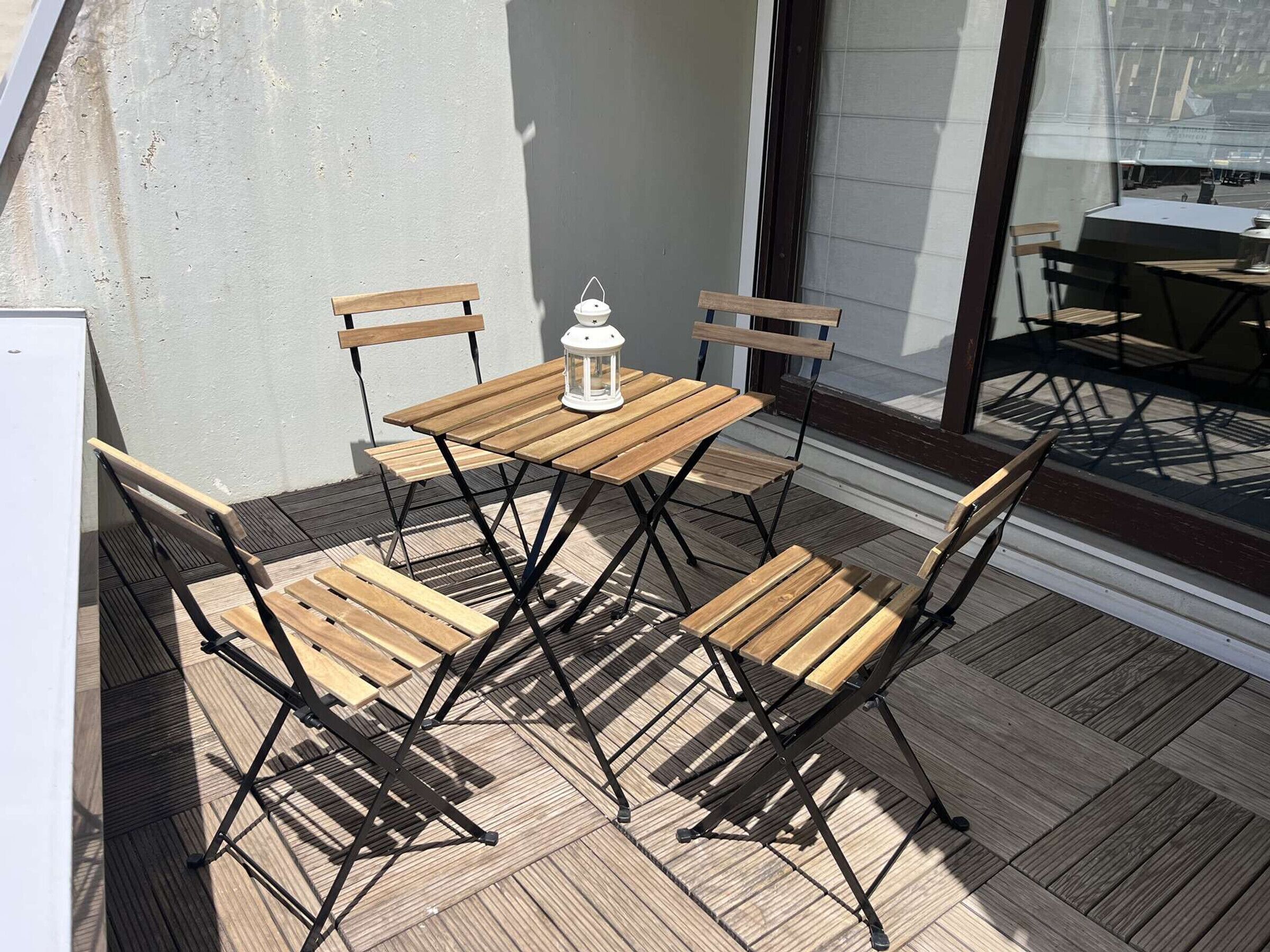 Eine Terrasse im Freien mit einem Holztisch und Stühlen, einem weißen Laternenlicht und Blick auf die Straße durch eine Glastür.