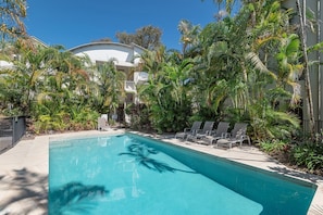 Una piscina con sillones de descanso, rodeada de plantas tropicales y un edificio al fondo.