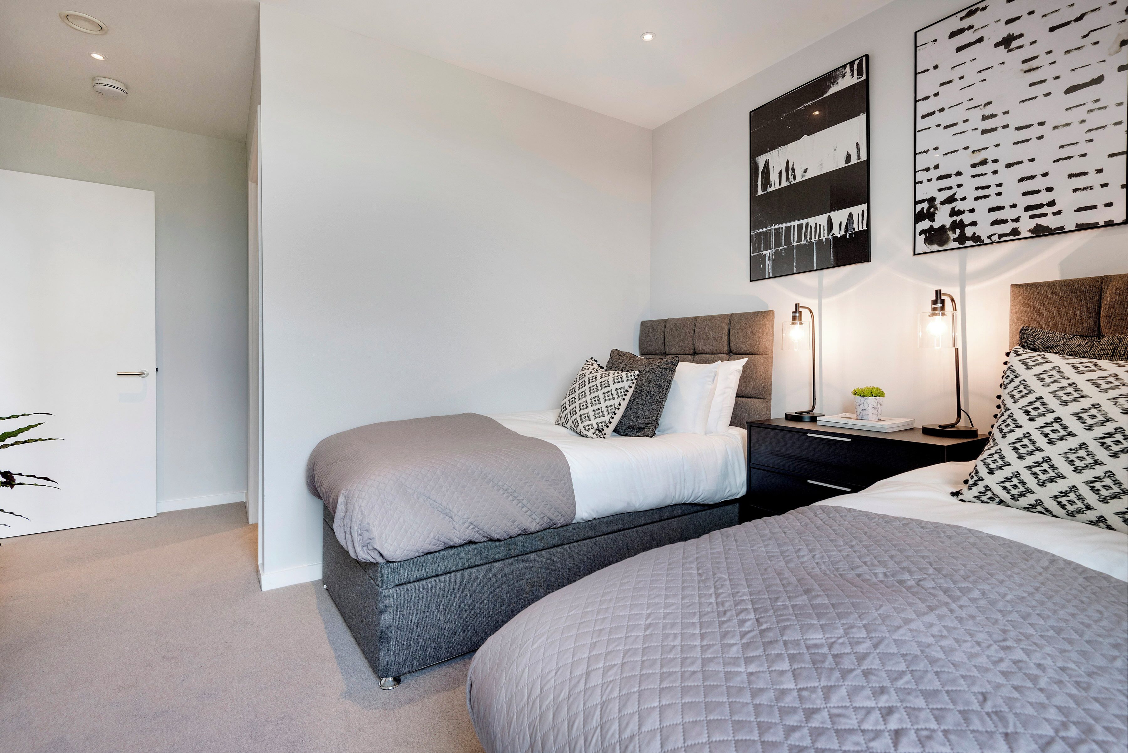 A modern bedroom with two beds, a dark headboard, and a bedside table with a lamp.