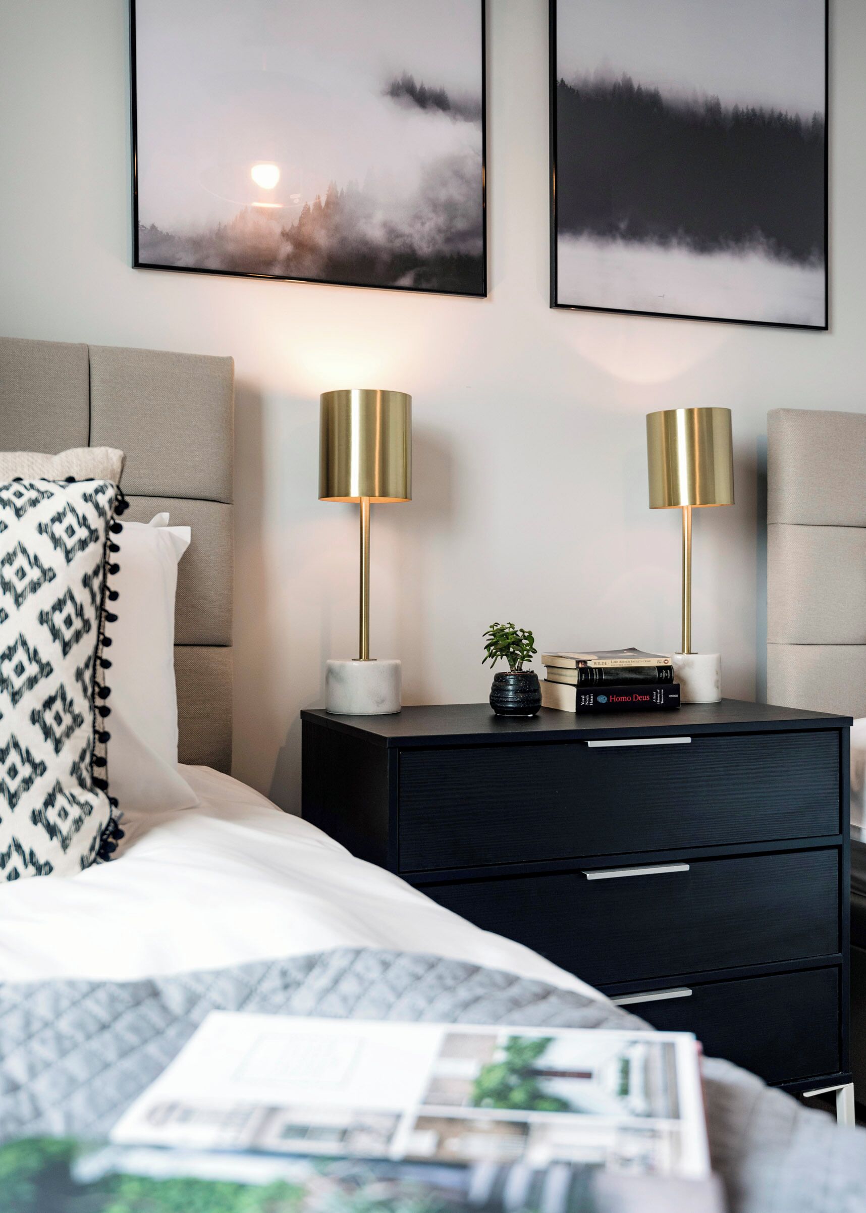 Une chambre à coucher bien rangée, avec un lit, deux lampes de chevet, une table de chevet avec des livres et une plante, et deux tableaux encadrés accrochés au mur.