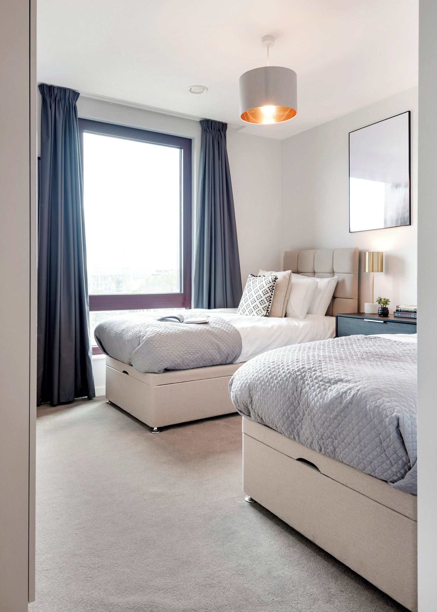 A modern bedroom with two beds, a large window with curtains, a bedside table with a lamp, and a ceiling light.