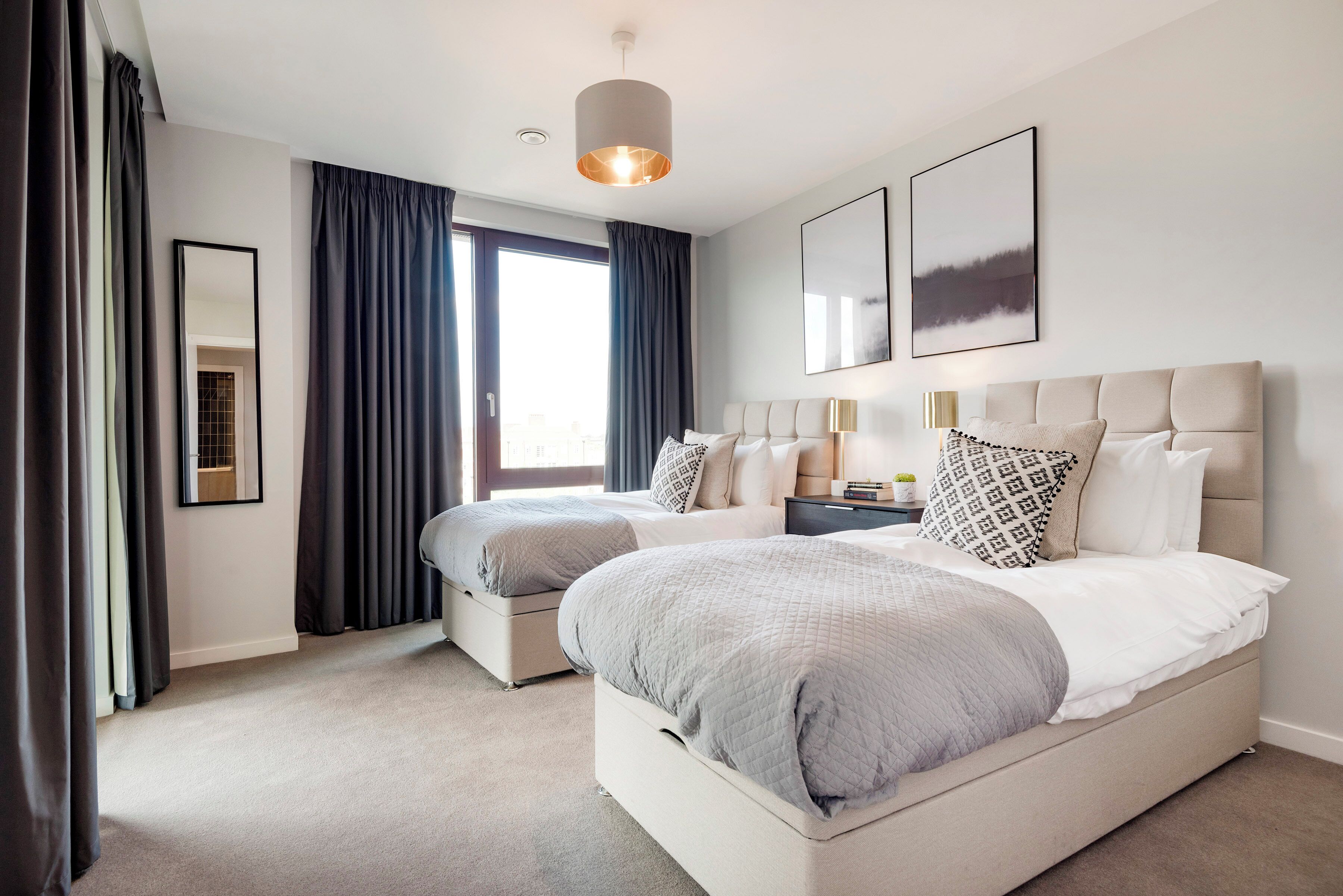 A hotel room with two beds, a large window with curtains, a hanging light fixture, and a framed picture on the wall.