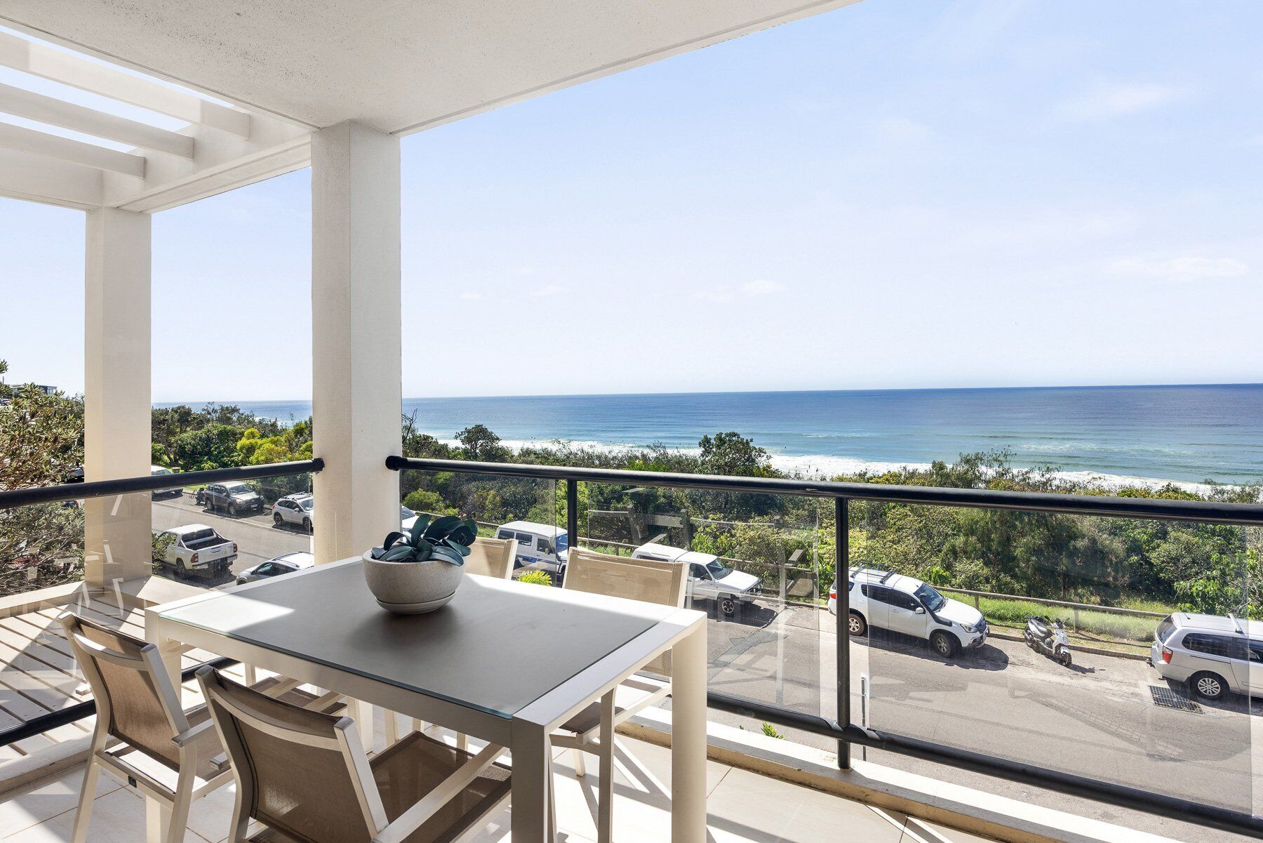 Un balcon avec une table et des chaises, donnant sur une plage et un parking.