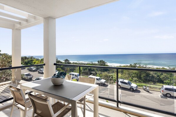 Un balcon avec une table et des chaises, donnant sur une plage et un parking.