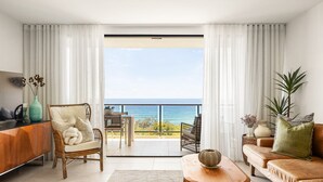 Un salon comprenant un canapé, un fauteuil, une table basse et un téléviseur. Il y a un balcon avec vue sur l’océan.